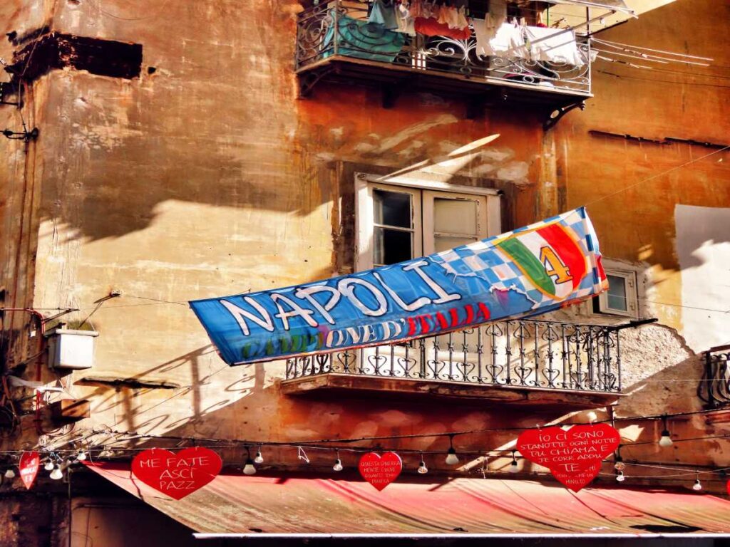 A Napoli il calcio fa parte della vita di tutti i giorni. Su un vecchio palazzo c’è uno striscione per festeggiare lo scudetto del Napoli. Intorno ci sono i balconi con i panni stesi e tanti cuori con i messaggi dei tifosi.
